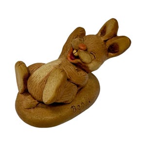 May include: A brown rabbit figurine in a reclining position, resting on an oval base. The rabbit has a wide, joyful expression with visible teeth, a small orange nose, and long ears. The name Benji is inscribed on the front of the base.