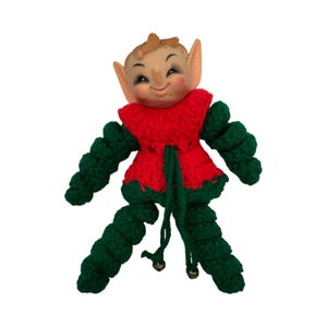 May include: A doll with a molded plastic head featuring pointed ears, painted facial features, and rosy cheeks. The body is crafted from red and dark green yarn, featuring a crocheted red bodice and dark green spiral-shaped arms and legs.