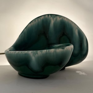 May include: A deep teal ceramic bowl with a smooth, glossy finish and a distinctive curved, asymmetrical shape. The glaze features a gradient effect, transitioning from a dark, saturated forest green at the base to a lighter, creamy white rim.