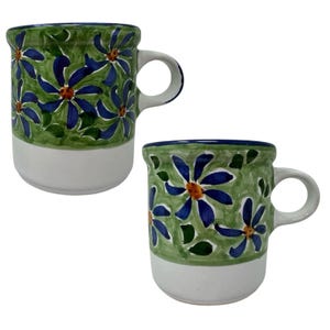 May include: Two ceramic mugs featuring a hand-painted design of blue flowers with orange centers on a green leafy background. The upper portion of each mug is decorated with this floral pattern, while the lower portion remains a solid white.