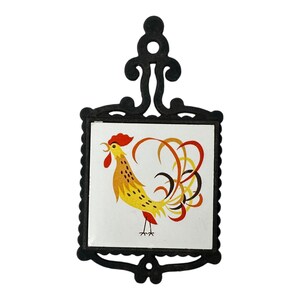 May include: A black cast iron trivet featuring a square white ceramic tile insert. The tile displays a stylized rooster illustration with yellow, orange, and red feathers, a red comb, and a long, flowing tail. The metal frame has a decorative handle.