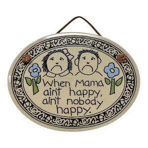 May include: An oval ceramic wall plaque with a cream-colored background and a black decorative border. It features hand-drawn illustrations of two sad-faced children and two blue flowers. The text reads: When Mama ain't happy, ain't nobody happy.