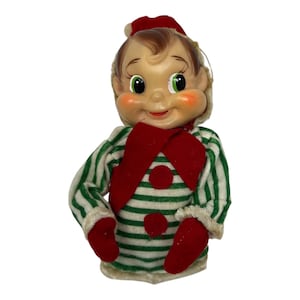 May include: A vintage-style elf doll with a molded plastic face, green eyes, and rosy cheeks. The doll wears a green and white striped long-sleeve shirt, a red collar, red mittens, and a red hat with a white trim, featuring two red circular buttons.