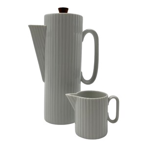 May include: A white porcelain coffee pot and a matching creamer pitcher, both featuring a vertical ribbed texture. The tall coffee pot has a large handle, a pointed spout, and a small brown wooden knob on the lid. The smaller creamer has a handle.