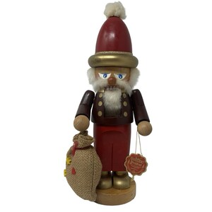 May include: A wooden nutcracker figure wearing a red hat with a white pom-pom and a gold brim. The figure has a white faux fur beard, a dark brown jacket with gold buttons, and red trousers. It holds a burlap sack and has a tag attached to its arm.