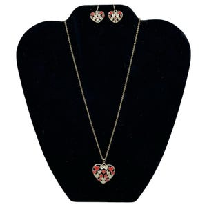 May include: A silver-toned necklace and earring set. The necklace features a heart-shaped pendant with red and white heart accents. The matching earrings also have heart designs. The jewelry is displayed on a black mannequin.