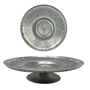 May include: A silver-toned metal pedestal tray featuring a circular base and a wide, shallow plate. The plate surface is decorated with an embossed floral pattern of roses and leaves, with a notched rim edge and a smooth, recessed center.