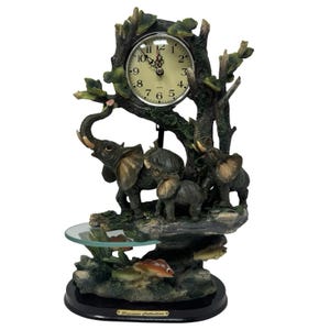 May include: A decorative table clock featuring a round face with black numerals set into a sculpted tree trunk base. Three grey elephants are positioned among the branches, with small fish figures below and a glass shelf. A plaque reads Precious Collection.