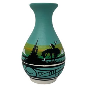 May include: A teal vase with a flared neck and a rounded body. The surface features a painted landscape with a black silhouette of a horse and rider, green and yellow gradients, white mountain peaks, and black geometric patterns at the base.
