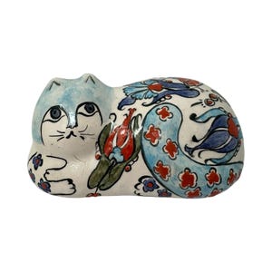 May include: A ceramic cat figurine in a reclining position, featuring a white base with hand-painted floral motifs in shades of blue, red, and green. The cat has a light blue face, dark blue eyes, and a tail decorated with stylized tulip patterns.