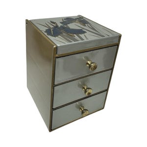 May include: A small chest of drawers with a metallic gold finish and three stacked drawers. Each drawer features a round gold knob. The top surface displays a painted illustration of dark blue iris flowers with green stems on a light gray background.
