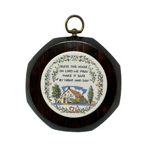 May include: A circular wall hanging featuring a dark wood frame with a brass ring at the top. The center displays a cross-stitch embroidery of a house with trees and the text: BLESS THIS HOUSE OH LORD WE PRAY MAKE IT SAFE BY NIGHT AND DAY.