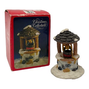 May include: A miniature wishing well figurine featuring a stone-textured base, a dark shingled roof with white snow accents, and a small bucket suspended by a rope. The item is displayed next to its red cardboard box labeled Christmas Collectable.