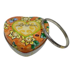 May include: A heart-shaped metal tin featuring a sunflower and bee pattern with the word Brighton printed on the lid, accompanied by a silver-toned metal bangle bracelet with an engraved scrollwork design along its outer surface.