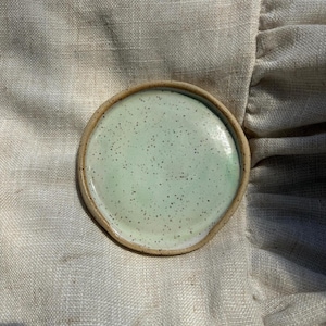May include: A round ceramic dish featuring a light mint green glaze with dark brown speckles throughout. The dish has a raised, unglazed tan rim that frames the interior surface, creating a distinct border around the speckled green centre.
