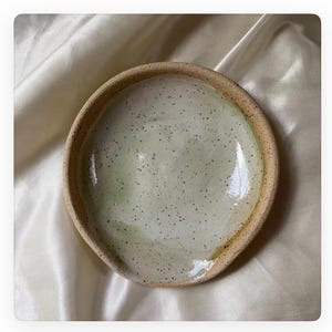 May include: A speckled, off-white ceramic bowl with a slightly uneven rim. The bowl has a light green glaze and a speckled finish.