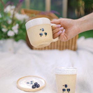 Peut inclure: Un mug en céramique avec anse, un gobelet assorti et une petite assiette circulaire, tous dotés d'une finition crème mouchetée et de motifs de baies bleues peints à la main. Le mug présente un bord blanc, tandis que le gobelet et l'assiette sont assortis.
