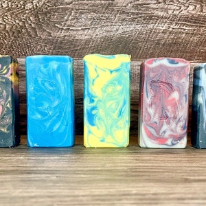 May include: Five bars of handmade soap with swirling patterns in shades of black, blue, yellow, red, and white. The soap bars are arranged in a row on a wooden surface.