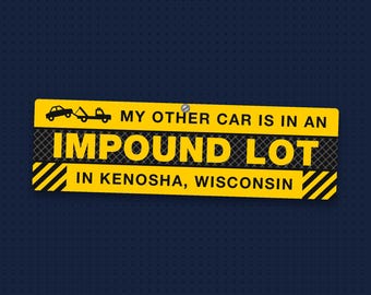 May include: A rectangular yellow and black metal sign featuring a graphic of a tow truck and the text: MY OTHER CAR IS IN AN IMPOUND LOT IN KENOSHA, WISCONSIN. The sign includes a small screw hole at the top centre and diagonal hazard stripes.