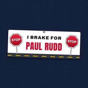 May include: A rectangular white bumper sticker featuring the black text I BRAKE FOR and the red text PAUL RUDD. Two red octagonal stop signs are positioned on either side of the text, above a black road graphic with yellow dashed lines.