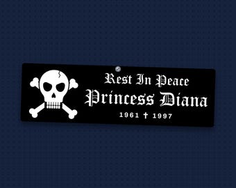 May include: A rectangular black sign featuring a white skull and crossbones graphic on the left side. The text reads Rest In Peace Princess Diana 1961 † 1997 in a white gothic font. A small silver screw is visible at the top center edge.