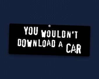 May include: A rectangular black sign featuring the white text "YOU WOULDN'T DOWNLOAD A CAR" in a distressed, hand-lettered font. The sign is secured at the top center with a single small silver screw, creating a simple, bold visual display.
