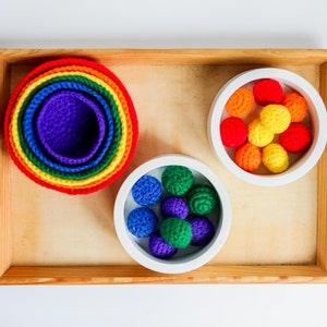 Puede incluir: Una bandeja de madera con tres cuencos blancos. Los cuencos contienen bolas de crochet de colores. Un cuenco tiene bolas de colores arcoíris, otro tiene bolas azules y verdes, y el último tiene un juego de cuencos anidados de colores arcoíris.