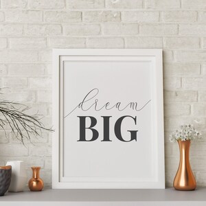 Puede incluir: Impresión enmarcada en blanco con el texto "dream BIG" en letras negras sobre fondo blanco. La impresión se muestra en una pared de ladrillo blanco con un jarrón gris, un jarrón blanco, un tazón gris y un jarrón de cobre con flores blancas.