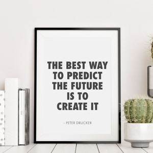 May include: A framed print with the quote "THE BEST WAY TO PREDICT THE FUTURE IS TO CREATE IT" by Peter Drucker. The black frame sits on a white surface with books and potted cacti.