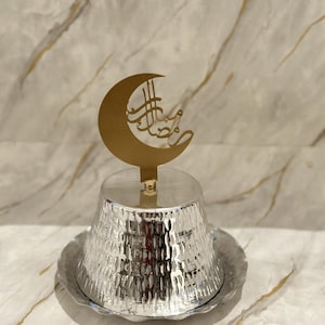 May include: A silver serving dish with a hammered finish and a gold crescent moon design on top. The crescent moon has Arabic calligraphy.