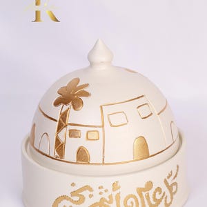 Könnte beinhalten: Weißer, kuppelförmiger Räucherofen aus Keramik mit einem goldenen Design aus einem Palmenbaum und Gebäuden. Der Sockel des Räucherofens trägt den arabischen Schriftzug "Ramadan Kareem" in Gold.