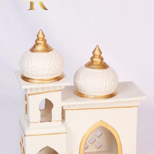 Può includere: Portacandele in ceramica bianca a forma di moschea con dettagli dorati. Il portacandele ha due cupole e tagli intricati.