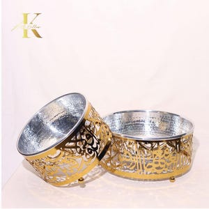 May include: Two decorative metal bowls. The bowls have a hammered silver interior and a gold-coloured filigree exterior. The filigree design features an intricate pattern. The bowls are round and sit on small feet. The logo "K" is in the upper left corner.
