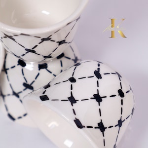 Puede incluir: Un juego de cuatro hueveras de cerámica blanca con un patrón geométrico en blanco y negro. Las hueveras están apiladas una encima de la otra.