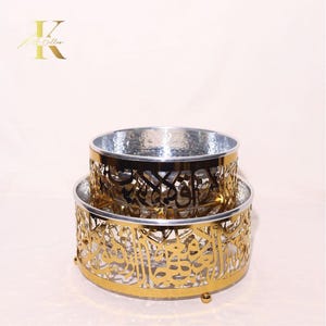 May include: Two-tiered decorative bowls with intricate gold filigree patterns and silver interiors. The bowls feature Arabic calligraphy designs. The lower bowl has a round base with small feet. The logo "K" is in the upper left corner.