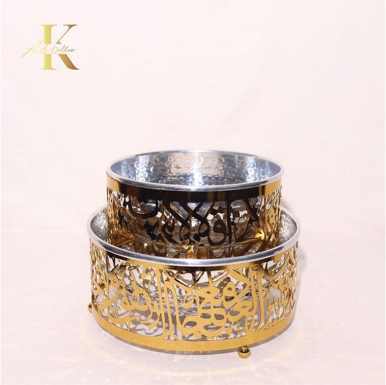 May include: Two-tiered decorative bowls with intricate gold filigree patterns and silver interiors. The bowls feature Arabic calligraphy designs. The lower bowl has a round base with small feet. The logo "K" is in the upper left corner.