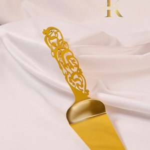 Könnte beinhalten: Goldener Kuchenspachtel mit einem verzierten, dekorativen Griff. Der Spachtel hat eine breite, flache Klinge.