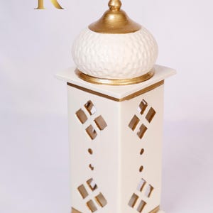 May include: White ceramic incense burner with a gold-painted dome top and intricate cut-out designs. The burner has a square base and a tall, rectangular body.