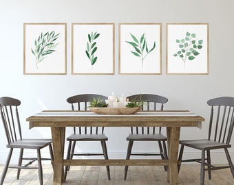 May include: Four framed watercolor paintings of green leaves hang on a white wall above a wooden dining table with four chairs. The table is set with a white tablecloth and a wooden bowl filled with greenery.