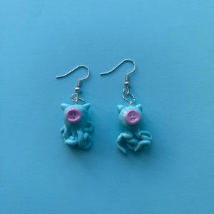 May include: A pair of dangle earrings featuring light blue octopus charms with small pointed ears and a single pink four-hole button centered on the head. The charms are attached to silver-toned metal fishhook ear wires with jump rings.