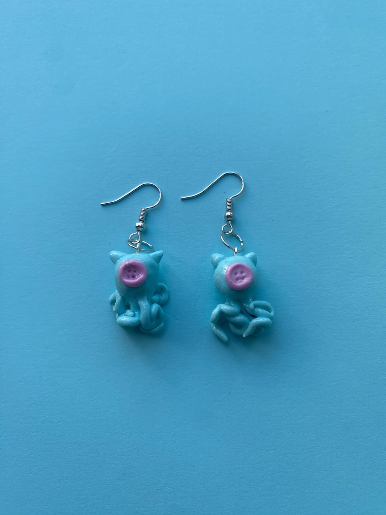 May include: A pair of dangle earrings featuring light blue octopus charms with small pointed ears and a single pink four-hole button centered on the head. The charms are attached to silver-toned metal fishhook ear wires with jump rings.