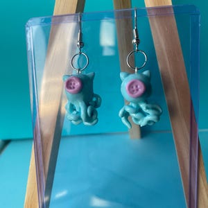 May include: A pair of dangle earrings featuring light blue octopus-shaped charms. Each octopus has a small, round, light purple button attached to its face, serving as a nose. The charms hang from silver-toned metal earring hooks with jump rings.
