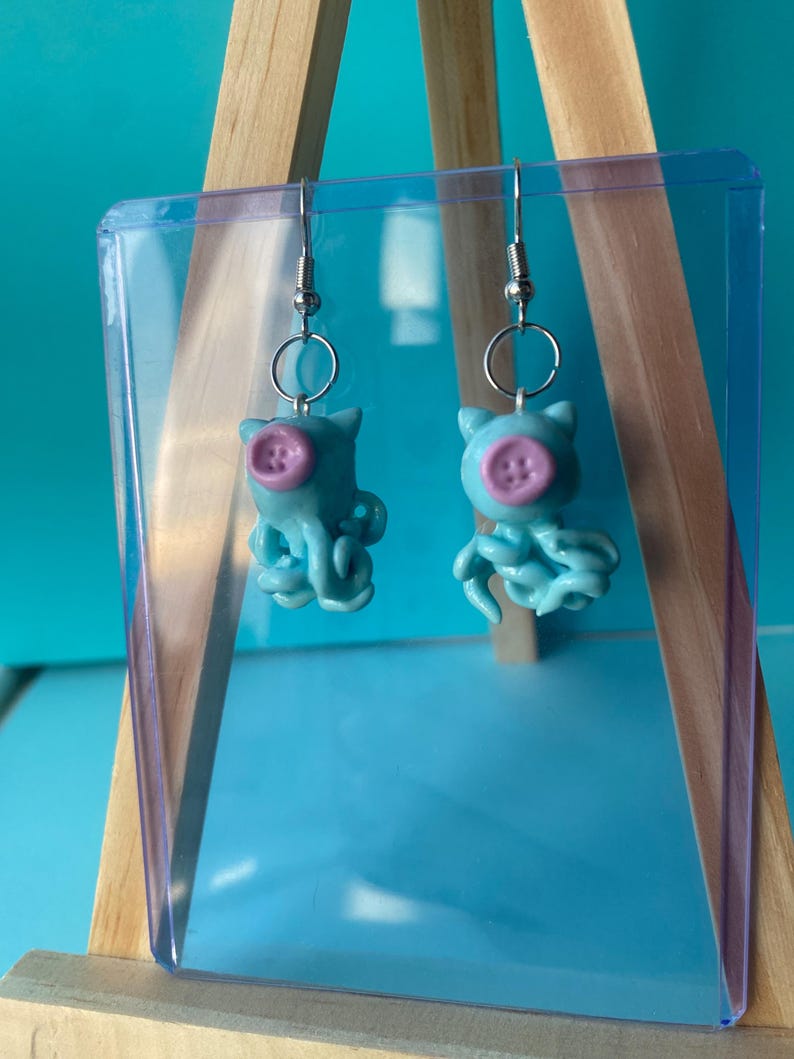 May include: A pair of dangle earrings featuring light blue octopus-shaped charms. Each octopus has a small, round, light purple button attached to its face, serving as a nose. The charms hang from silver-toned metal earring hooks with jump rings.
