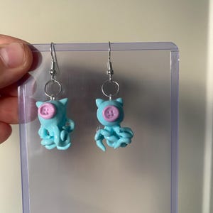 May include: A pair of dangle earrings featuring light blue octopus-shaped charms with small pink buttons attached to the center of their heads. The charms are suspended from silver-toned metal fishhook wires by small circular jump rings.