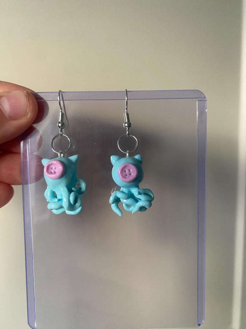 May include: A pair of dangle earrings featuring light blue octopus-shaped charms with small pink buttons attached to the center of their heads. The charms are suspended from silver-toned metal fishhook wires by small circular jump rings.