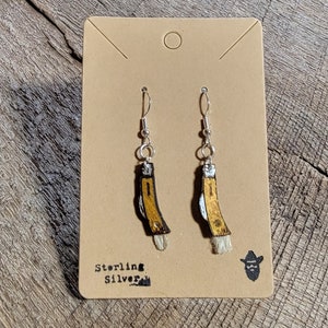 May include: A pair of sterling silver earrings featuring a miniature pocket knife design. The earrings have a silver hook and a small, tan-colored knife with a yellow handle and a white tassel at the end. The card reads "Sterling Silver."