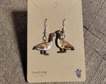 Op de afbeelding: Een paar sterling zilveren oorbellen met een houten uitsnijding van een Canadese gans. De oorbellen worden getoond op een bruine kaart met de tekst "Sterling Silver" en een klein cartoonbeeld van een man met een cowboyhoed.