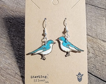 May include: A pair of sterling silver earrings featuring a blue and white bird design. The earrings are on a brown card with the text "Sterling Silver" and a small illustration of a bearded man wearing a hat.