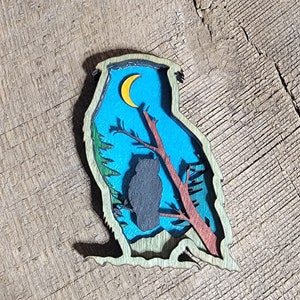 May include: A layered wooden owl-shaped art piece. The design features a silhouette of an owl perched on a branch, a yellow crescent moon, and a blue sky background. The outer frame is green, and the owl is dark gray.