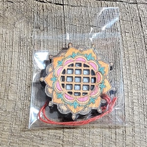 Puede incluir: Un adorno de madera con forma de mandala con un diseño rosa, naranja y verde. El mandala tiene un patrón de cuadrícula en el centro y está colgado de una cuerda roja.
